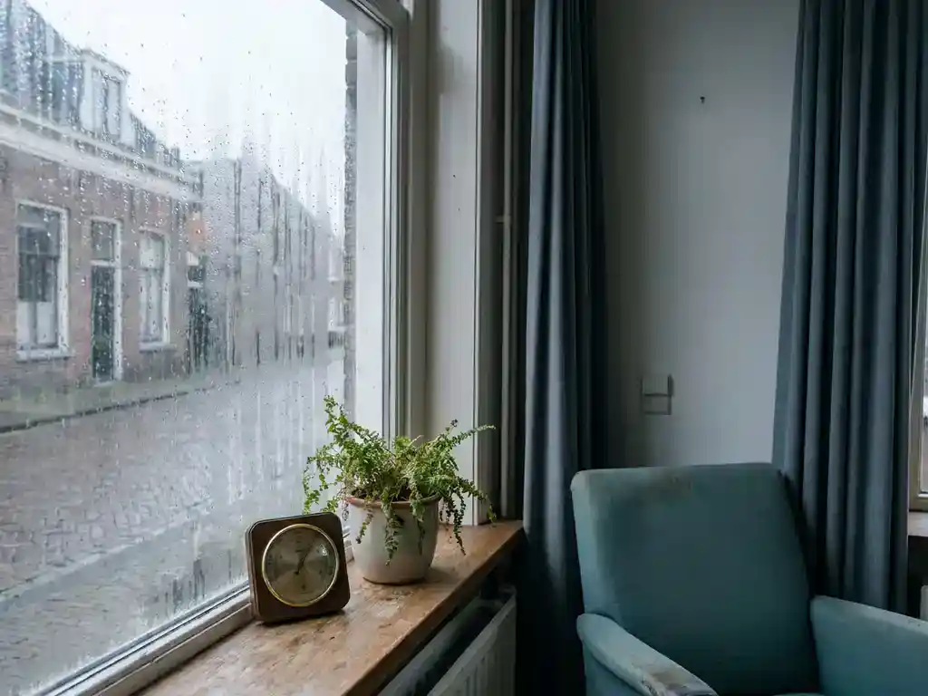 Beslagen raam in Nederlandse woning met condenswater op glas, hygrometer op vensterbank toont hoge luchtvochtigheid, grijs daglicht.