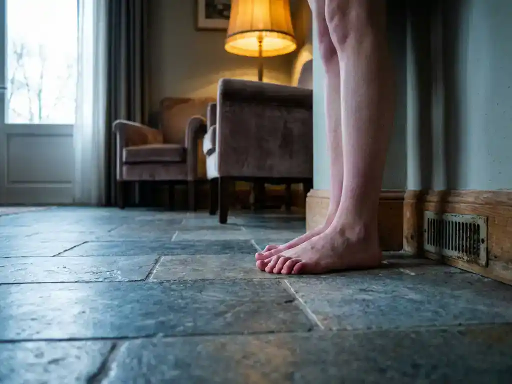 Blote voeten op koude stenen vloertegels in Nederlandse woonkamer, tenen opgetrokken tegen de kou, kruipruimterooster zichtbaar bij de plint.