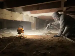 Installateur spuit isolatieschuim onder houten vloerbalken in een kruipruimte, verlicht door warm werklicht.