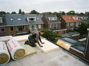 Dakspecialist die isolatieplaten aanbrengt op een plat betonnen dak, met minerale wolrollen en een Nederlandse woonwijk op de achtergrond.
