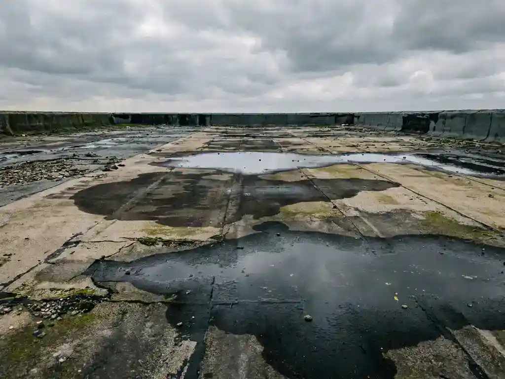 Verweerd plat dak met waterplassen en donkere vochtplekken op verouderd dakmembraan onder bewolkte Nederlandse lucht.